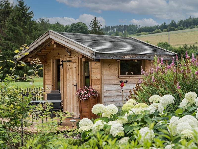 Eine kleine Holzhütte aus der örtlichen Holzhandlung mit schrägem Dach ist umgeben von blühenden Blumen und Grünzeug in einer ländlichen Gartenanlage unter einem teilweise bewölkten Himmel.